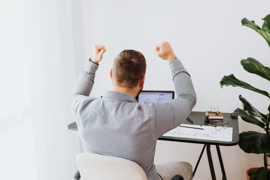 Back view of a man cheering with arms raised while sitting at a desk.