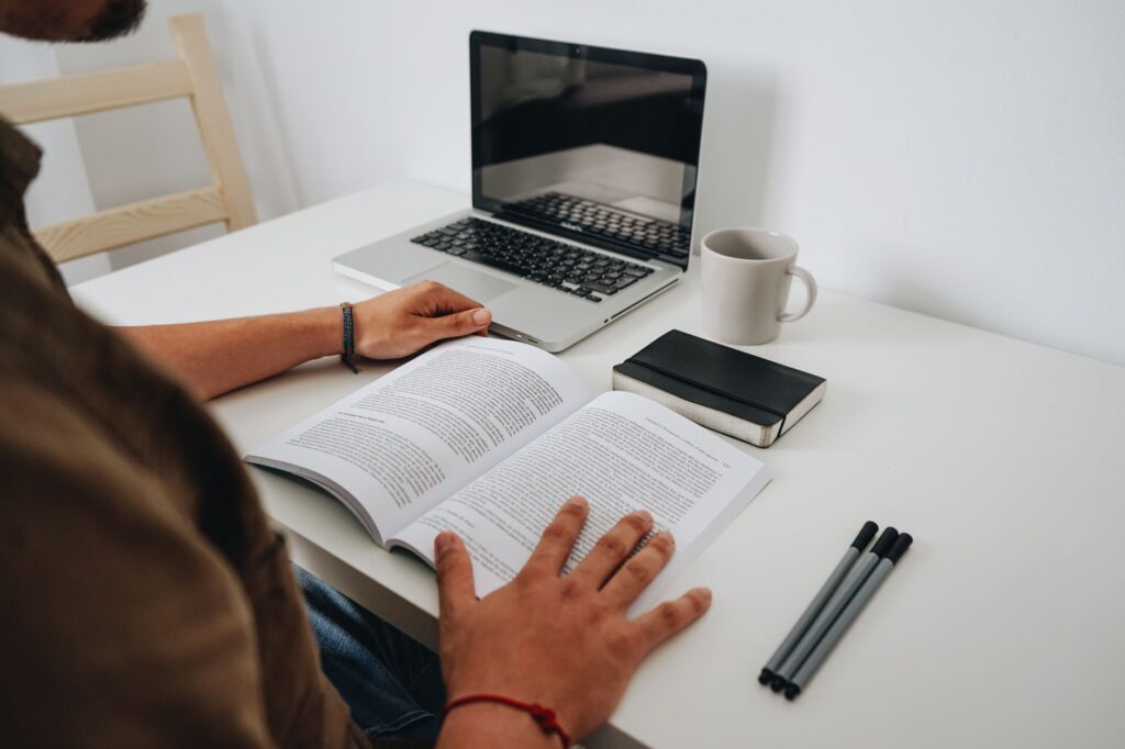 A person reading at a minimalist home office with a laptop, coffee mug, and book.