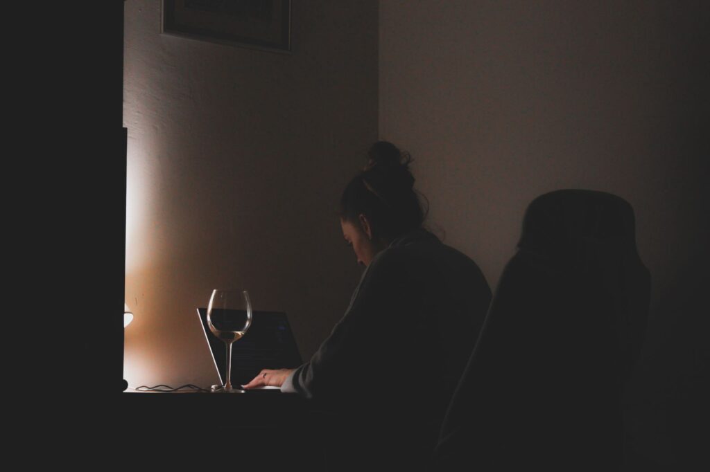 A woman working on a laptop with a glass of wine in a dimly lit room.